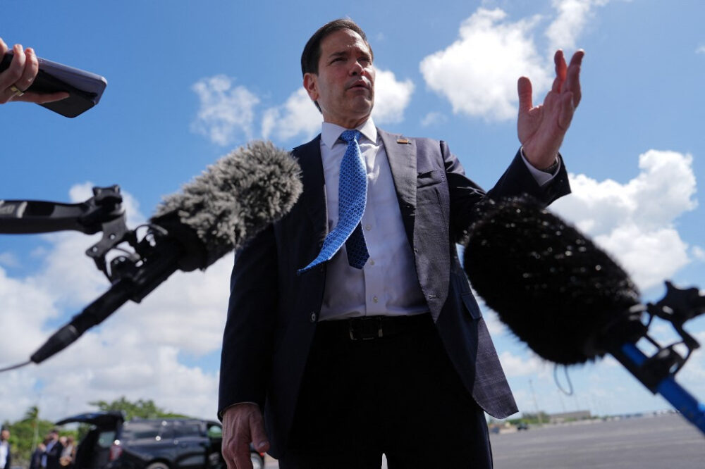 6Lcx15CYa7vB-sEWBJF1lN - MultiPol 360 Politician in a navy suit speaks to reporters, microphones in front, blue sky behind him.