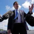 Politician in a navy suit speaks to reporters, microphones in front, blue sky behind him.