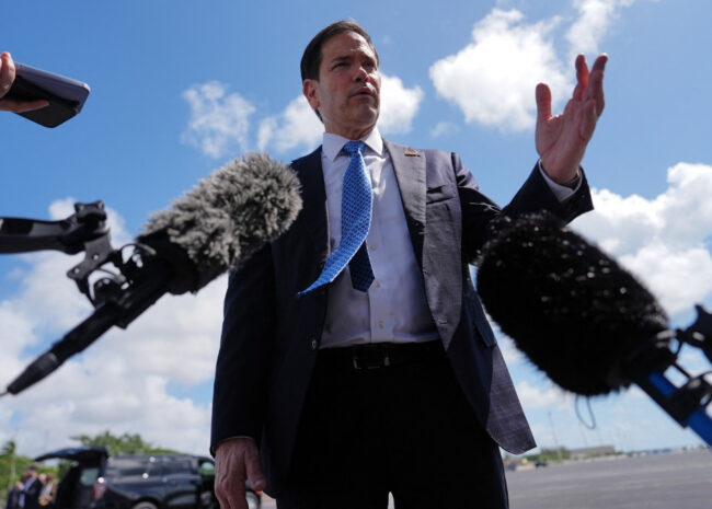Politician in a navy suit speaks to reporters, microphones in front, blue sky behind him.