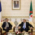 Two men in suits sit on cream armchairs in a formal meeting room with EU, French, and Algerian flags behind them.