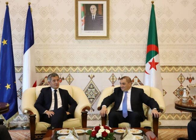 Two men in suits sit on cream armchairs in a formal meeting room with EU, French, and Algerian flags behind them.