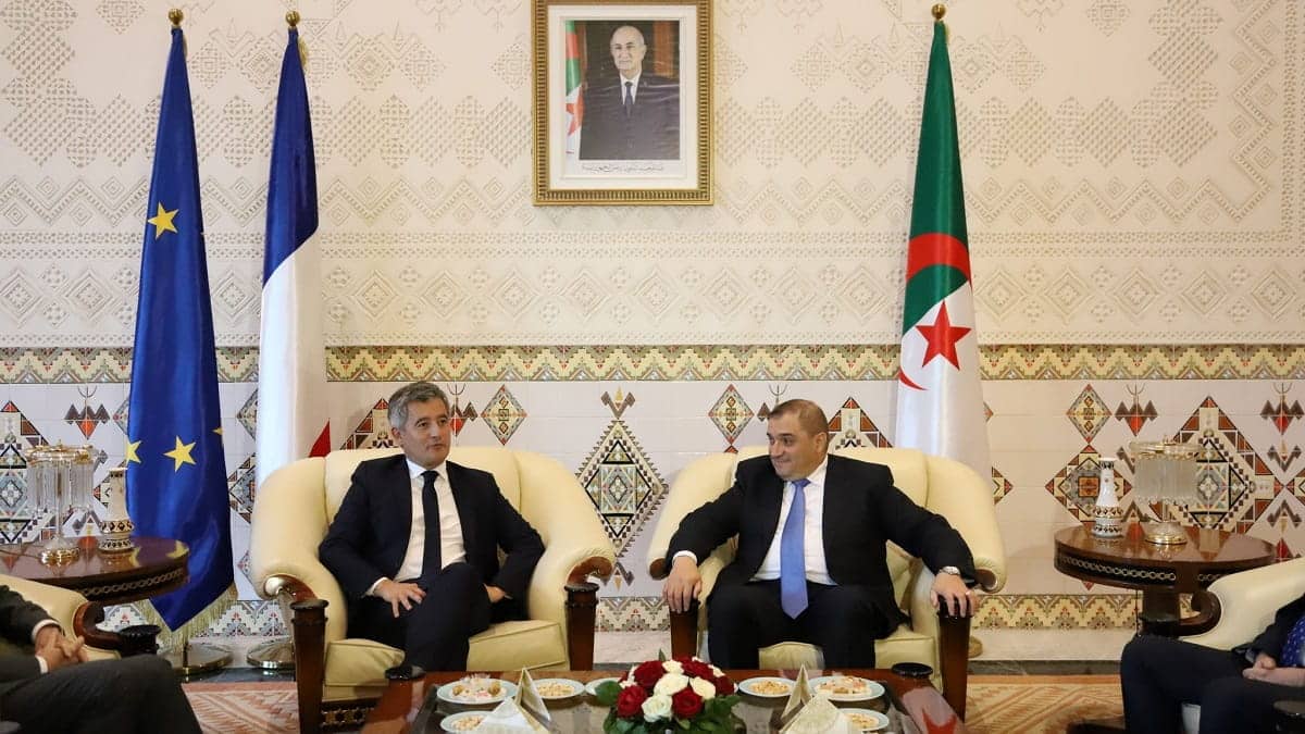 Two men in suits sit on cream armchairs in a formal meeting room with EU, French, and Algerian flags behind them.