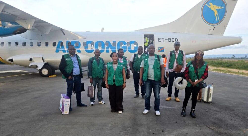 Group of airline staff in green vests posing on the tarmac in front of a blue-and-white Air Congo airplane, tail logo visible.