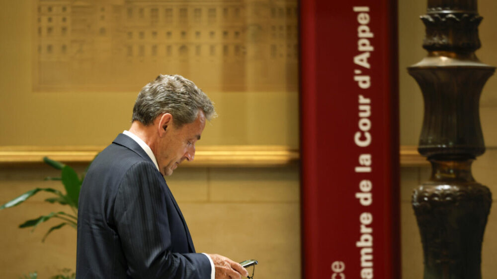 Man in a dark suit looks at his phone in a courthouse lobby, beside a red vertical sign reading 'Cour d'appel' and a tall ornate column.