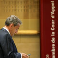 Man in a dark suit looks at his phone in a courthouse lobby, beside a red vertical sign reading 'Cour d'appel' and a tall ornate column.