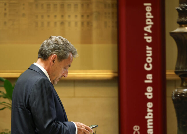 Man in a dark suit looks at his phone in a courthouse lobby, beside a red vertical sign reading 'Cour d'appel' and a tall ornate column.