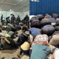 Large group of people kneeling and bowing in prayer inside a temporary shelter or tent, crowded on the floor.