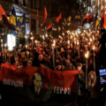 Night torchlit march with a large crowd carrying flags and a red banner with a portrait.