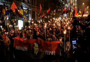 Night torchlit march with a large crowd carrying flags and a red banner with a portrait.
