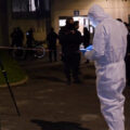 Nighttime scene of a crime investigation with officers behind red-and-white tape and a person in white protective suit examining the area near an entrance. A tripod and equipment stand in the foreground.