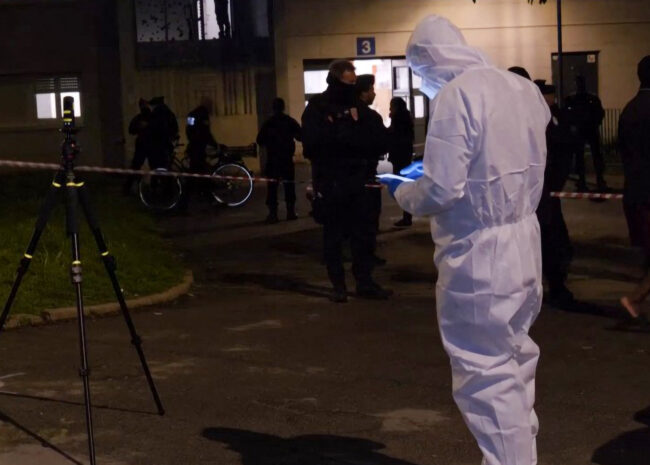 Nighttime scene of a crime investigation with officers behind red-and-white tape and a person in white protective suit examining the area near an entrance. A tripod and equipment stand in the foreground.