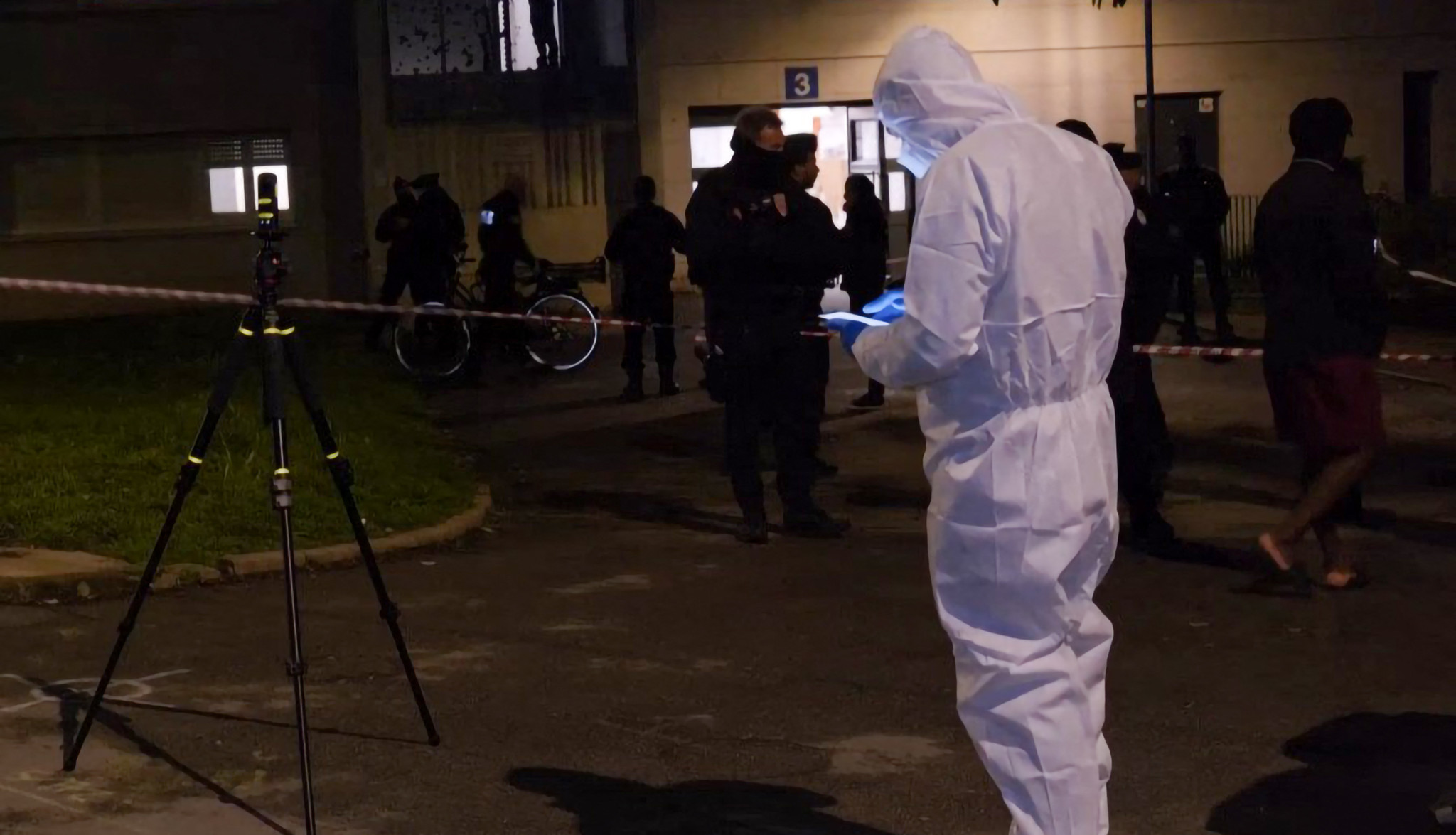 Nighttime scene of a crime investigation with officers behind red-and-white tape and a person in white protective suit examining the area near an entrance. A tripod and equipment stand in the foreground.