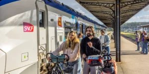 People with bicycles and helmets gather at a platform beside a white SNCF train with a Région Normandie emblem, smiling and ready to board.