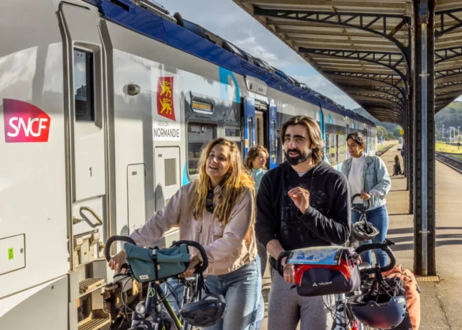 People with bicycles and helmets gather at a platform beside a white SNCF train with a Région Normandie emblem, smiling and ready to board.