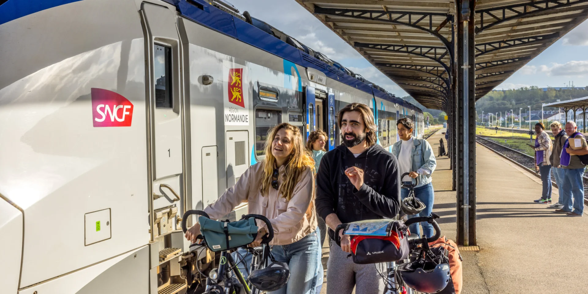 People with bicycles and helmets gather at a platform beside a white SNCF train with a Région Normandie emblem, smiling and ready to board.