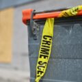 Grey dumpster with bright yellow crime scene tape draped across the top, in an industrial area.
