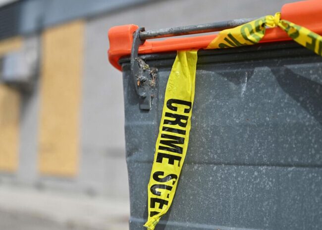 Grey dumpster with bright yellow crime scene tape draped across the top, in an industrial area.
