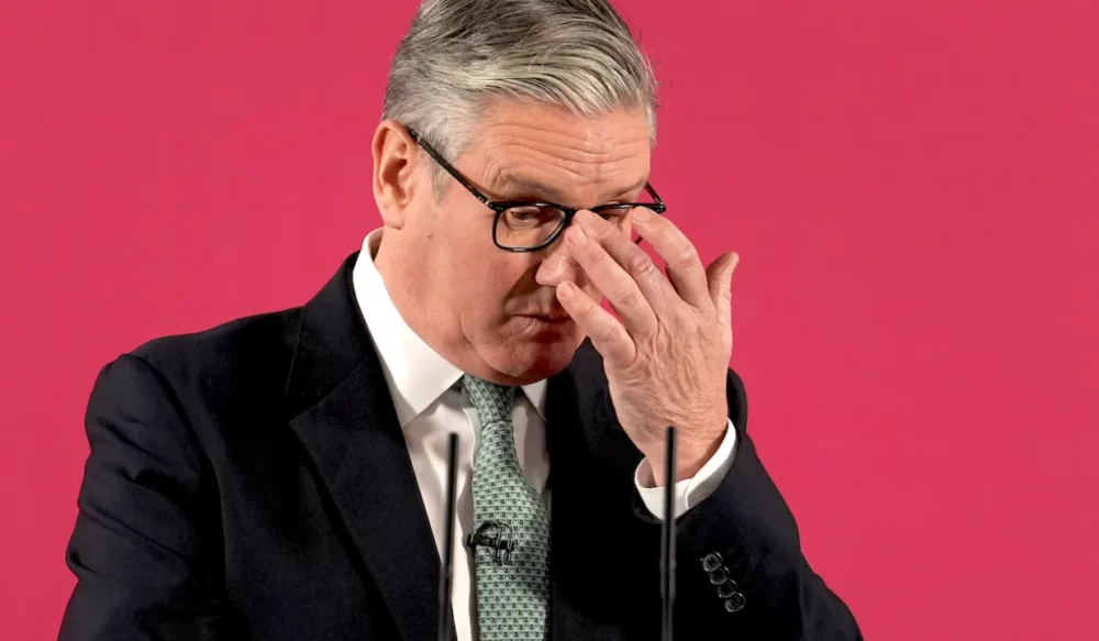 Man in a dark suit and green tie adjusts his glasses against a bright pink background, with a microphone in front.