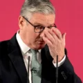 Man in a dark suit and green tie adjusts his glasses against a bright pink background, with a microphone in front.