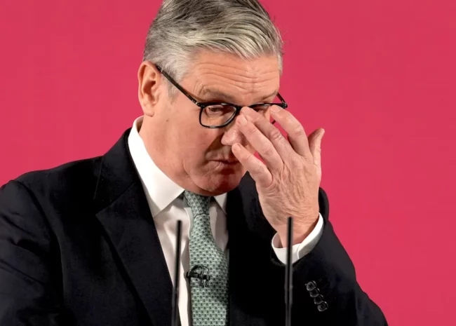Man in a dark suit and green tie adjusts his glasses against a bright pink background, with a microphone in front.