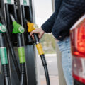 Close-up of gas pump dispensers with green and yellow nozzles; a person inserts a yellow nozzle into a car.
