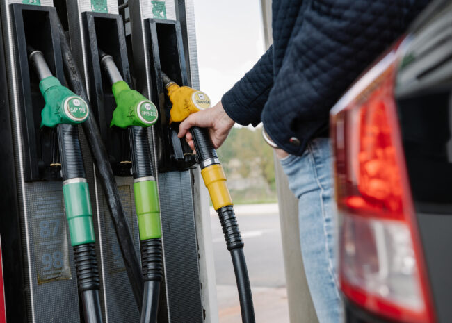 Close-up of gas pump dispensers with green and yellow nozzles; a person inserts a yellow nozzle into a car.