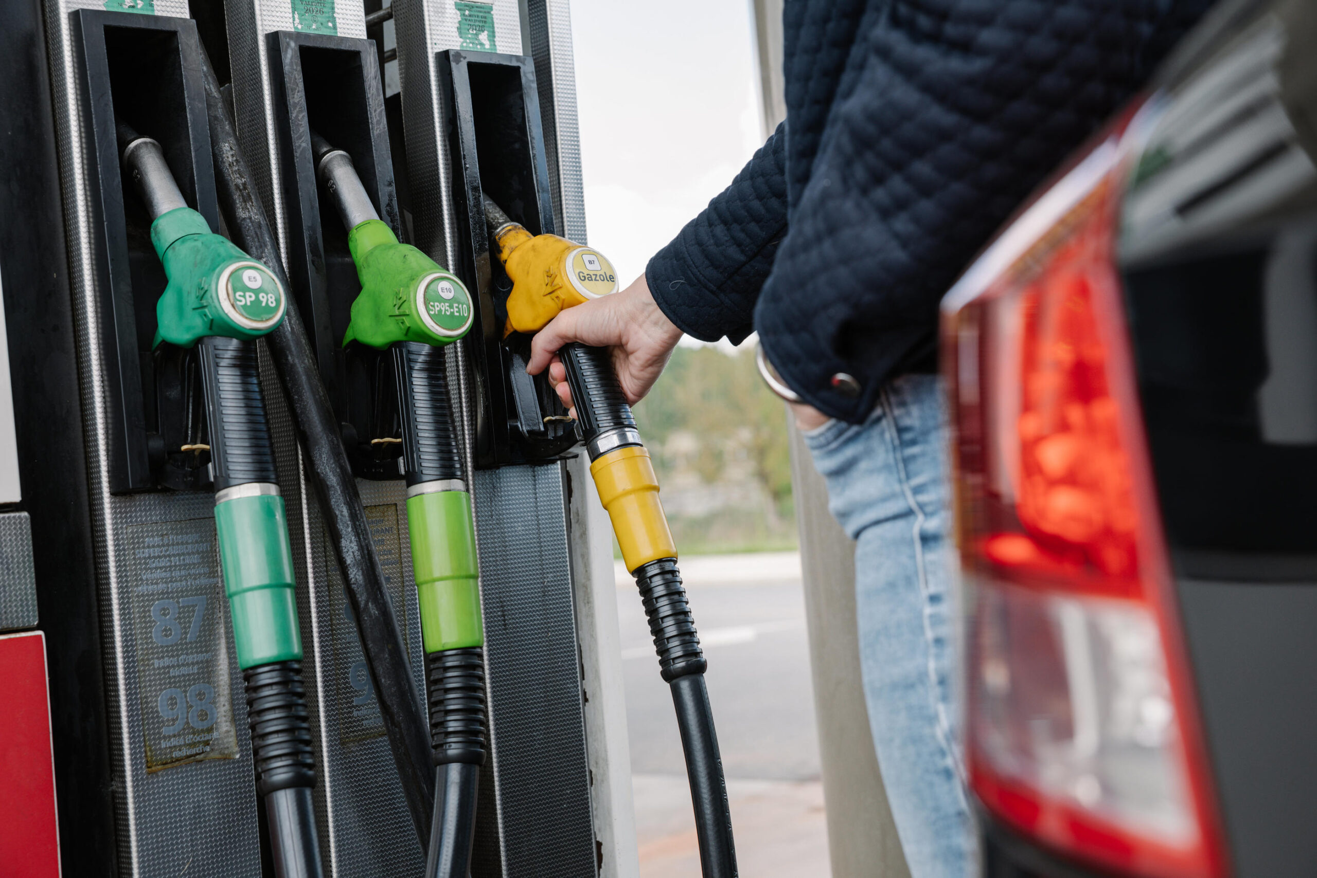 Close-up of gas pump dispensers with green and yellow nozzles; a person inserts a yellow nozzle into a car.