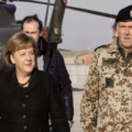 A woman in a black coat walks beside a camouflaged uniformed officer on a tarmac near a helicopter.