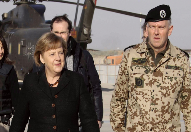 A woman in a black coat walks beside a camouflaged uniformed officer on a tarmac near a helicopter.