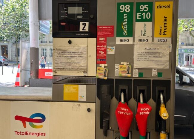Gas pump at a TotalEnergies station with multiple fuel options (98, 95 E10, diesel) and red 'hors service' hose covers; urban street in background.