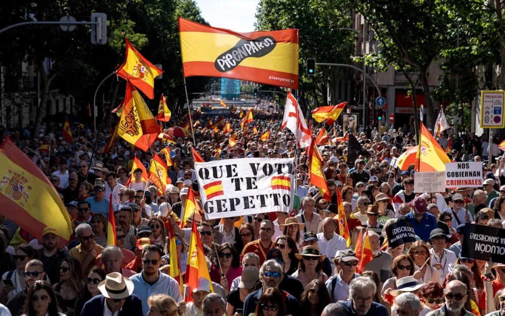 Large street protest with many people waving Spanish flags and banners, including a central sign that reads 'QUE VOTE TE GEPETTO'.