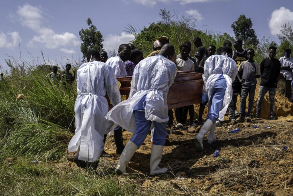 field_media_image-3015637-7baa01fe4ad0ca2f989b49fe98f0d7a980ecf20b - MultiPol 360 Mourners in white protective suits carry a wooden coffin through a dirt path as a crowd watches at an outdoor burial site.
