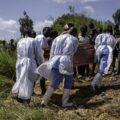Mourners in white protective suits carry a wooden coffin through a dirt path as a crowd watches at an outdoor burial site.