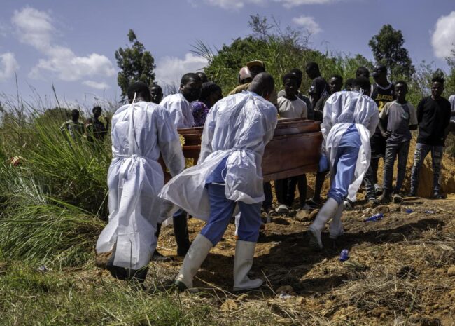 Mourners in white protective suits carry a wooden coffin through a dirt path as a crowd watches at an outdoor burial site.