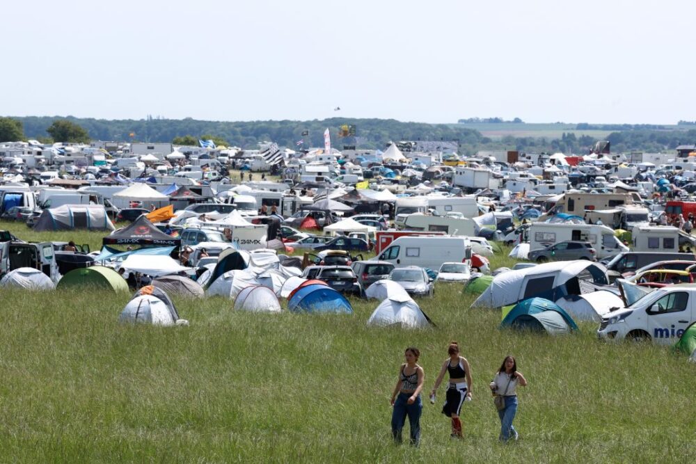 Wide campsite field crowded with tents, caravans, and cars at a festival; three people walk through the tall grass in the foreground.