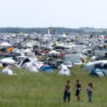 Wide campsite field crowded with tents, caravans, and cars at a festival; three people walk through the tall grass in the foreground.