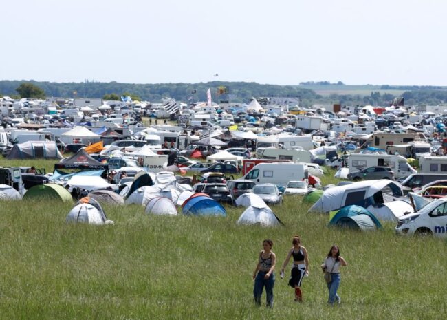 Wide campsite field crowded with tents, caravans, and cars at a festival; three people walk through the tall grass in the foreground.