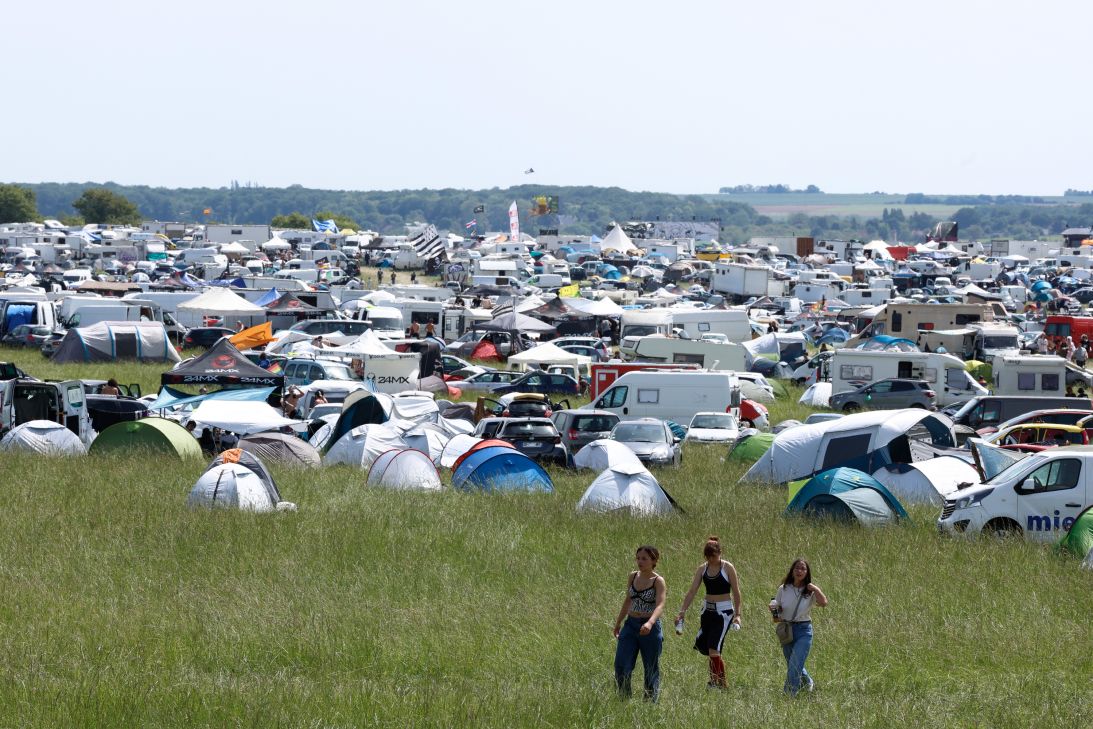 Wide campsite field crowded with tents, caravans, and cars at a festival; three people walk through the tall grass in the foreground.