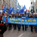 Crowd of protesters holding a blue banner reading 'BREXIT: Is It Worth It?' with EU flags; a bagpiper in the foreground on the left.