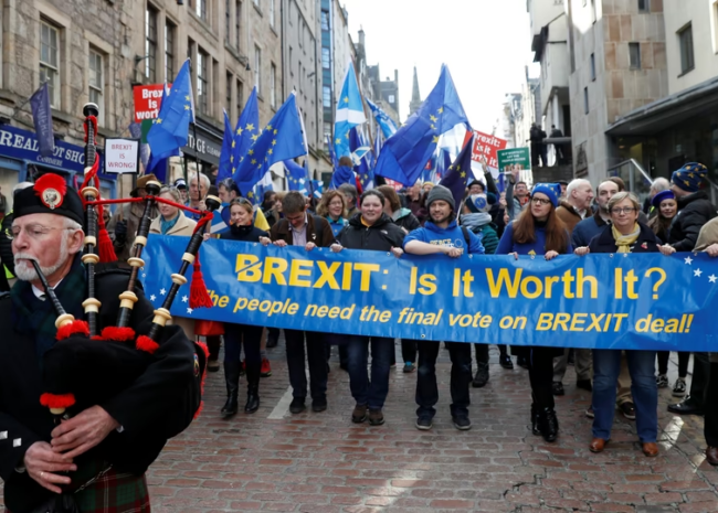 Crowd of protesters holding a blue banner reading 'BREXIT: Is It Worth It?' with EU flags; a bagpiper in the foreground on the left.
