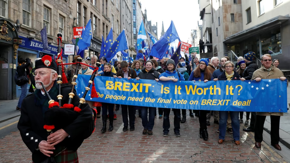 Crowd of protesters holding a blue banner reading 'BREXIT: Is It Worth It?' with EU flags; a bagpiper in the foreground on the left.