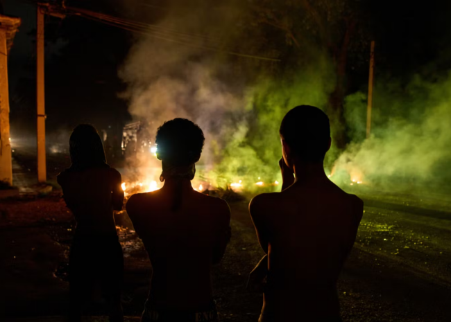 Three silhouetted figures with bare backs stand on a dark street at night, facing glowing embers and colored smoke.