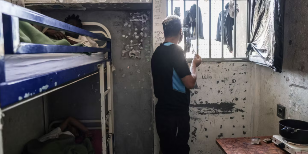 image_2026-05-28_12-59-54 - MultiPol 360 Man standing by a barred window in a cramped, peeling-wrothed dorm cell with bunk beds and a small kitchenette nearby.