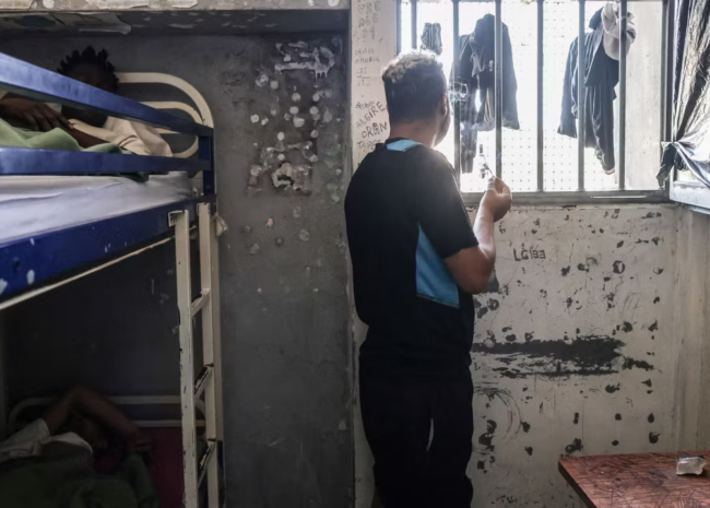 Man standing by a barred window in a cramped, peeling-wrothed dorm cell with bunk beds and a small kitchenette nearby.