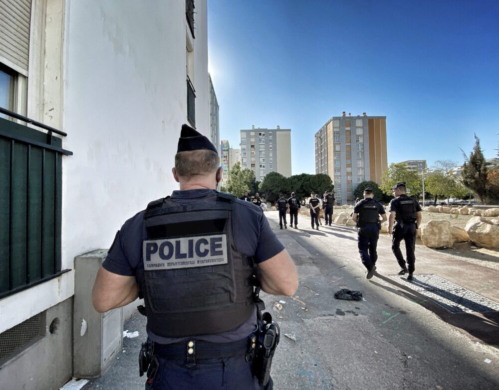 maxnewsspecialtwo361636-68f8a40c0b416556054584 - MultiPol 360 Back view of a police officer in tactical vest on an urban street with a group of officers in the distance