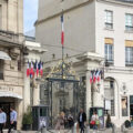 ornate wrought‑iron gate with gold accents and a French flag above, flanked by classic Parisian buildings and people walking on the street