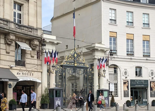 ornate wrought‑iron gate with gold accents and a French flag above, flanked by classic Parisian buildings and people walking on the street