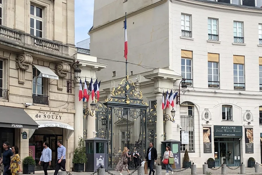 ornate wrought‑iron gate with gold accents and a French flag above, flanked by classic Parisian buildings and people walking on the street
