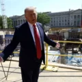 Politician in a dark suit and red tie speaks with arms outstretched on a construction site, with a large government-style building in the background and construction equipment around.