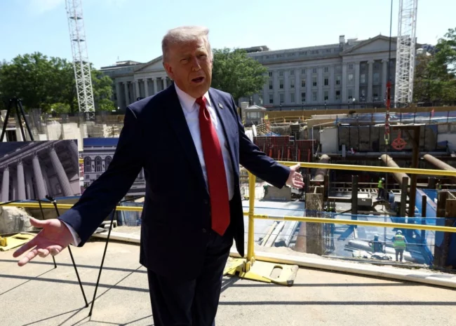Politician in a dark suit and red tie speaks with arms outstretched on a construction site, with a large government-style building in the background and construction equipment around.
